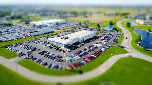 Aerial high-angle view of a bustling car dealership surrounded by parked cars in a green landscape.